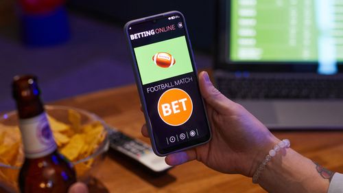 Person holding smartphone showing betting online app while drinking beer in living room, with laptop and snacks on wooden table