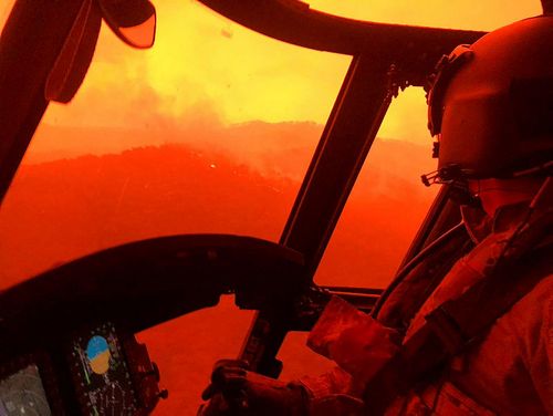 A photograph from inside a helicopter, as it flies above flames and smoke.