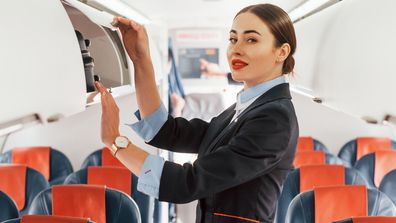 Young stewardess that is in formal black clothes is standing indoors in the plane.