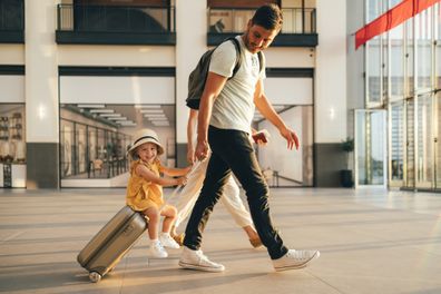 Cheerful husband and his anonymous wife walking with their little girl sitting on luggage at the airport.