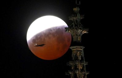 21 January 2019, North Rhine-Westphalia, Köln: The partly eclipsed so-called "blood moon" can be seen behind a tower of the Cologne Cathedral, which is also passed by an airplane.