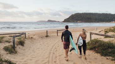 A rear-view shot of a young man and woman holding a surfing board on a beach in Sydney, Australia.