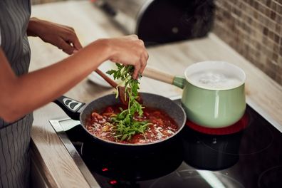 woman in apron standing next to stove, stirring tomato sauce and adding rocket. On stove is pot with boiling pasta.