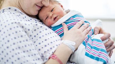 A caucasian mother in a hospital gown sits up in bed and holds her newborn baby to her chest