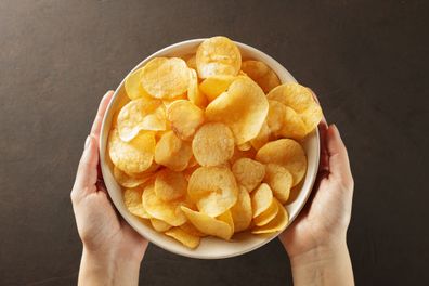 Hands giving bowl of potato chips on brown background. Concept of snack food. Top view.