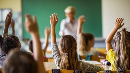 Rear view of large group of students raising their arms to answer the question on a class at elementary school.