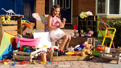 A little girl in a fairy costume sitting outside her house surrounded by used toys to sell in a yard sale.