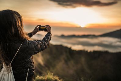 A woman stands alone at sunset high up on the beautiful Mount Batur and takes photos of the sunset with her smartphone.