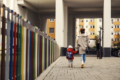 A young woman, mother of nanny, holding hands with little three year old boy with a schoolbag and taking him to kindergarten.