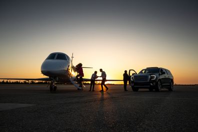 A young pilot welcomes passengers into private jet at dusk.