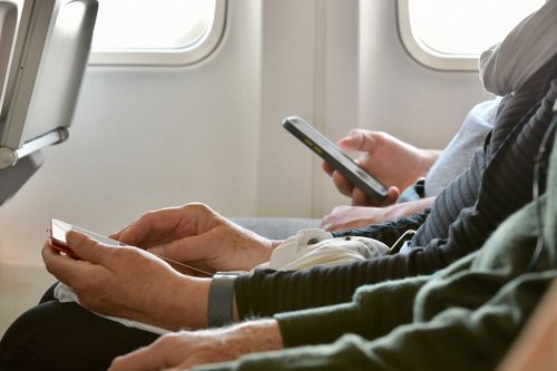 Long shot across laps of older person hands using cell phone.  Framed by seat back and clipped airplane windows.  