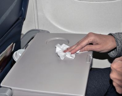 A woman sitting by a window seat is wiping down a germ laden dirty airplane tray with an antibacterial wipe illustrating the effort to stay healthy during air travel.