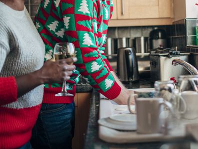 A woman holds a glass of white wine as she laughs with her husband in the kitchen whilst he washes the dishes.