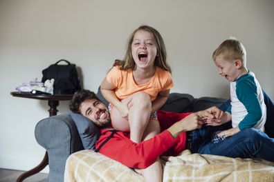 A shot of a mid adult male and his young niece and nephew wearing casual clothing. They are on a sofa in a living room and tickling the young girl.