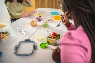 Young female employees are eating lunch together. They have healthy salads and dressings, and they eat out of plastic containers.