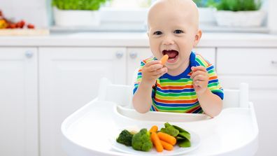 Cute baby eating vegetables in white kitchen. 