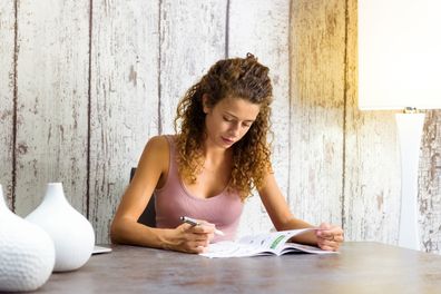 Young woman working on crossword puzzles at home in a magazine seated at a table.