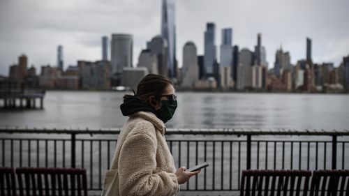 In this April 10, 2020 file photo a woman wearing a face mask sure to CVID-19 concerns walks along the Jersey City waterfront with the New York City skyline in the background. 