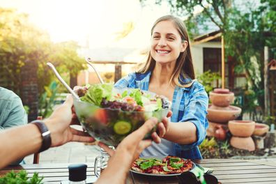 Shot of a young woman taking a bowl of salad from a friend while sitting around an outdoor table