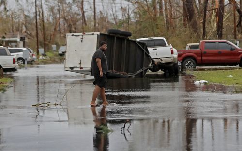 A man walks barefoot through the street with an overturned trailer in the distance.
