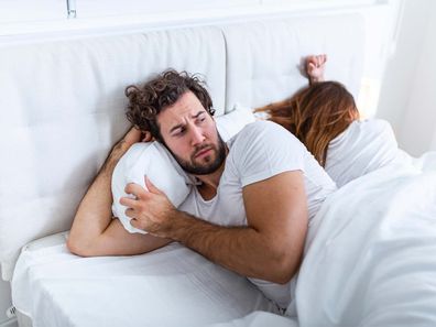 Man and woman lying in bed. The man is in front and looking suspicious of the wife, who is turned the other way, asleep.