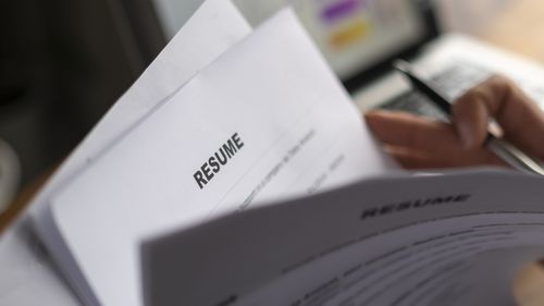 Uma foto sombria de mãos folheando currículos impressos com um laptop aberto ao fundo.
