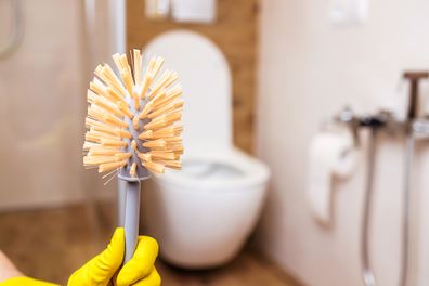 Close-up of a female hand in a yellow rubber cleaning glove holding a clean toilet brush. 