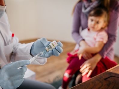 Doctor preparing vaccine with mum holding baby in background.