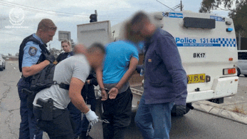 A man in a blue shirt with his face blurred being escorted into a police car in handcuffs.