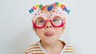 Cute little toddler boy in happy birthday glasses and party hat sticking his tongue out over white.