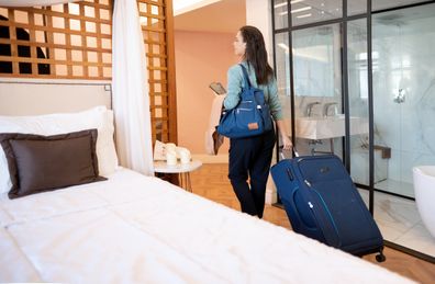Rear view of a young woman pulling her wheeled suitcase while leaving a hotel room to checkout