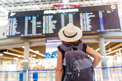 A woman who wears a straw hat and a backpack is stopped in front of a great airport exhibition. She is waiting for her flight, possibly at an airport.