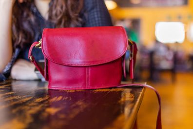 Cute red handbag on rustic-looking restaurant table with blurry background. Woman sitting at vintage table behind pretty purse with long straps. Fashion and accessories