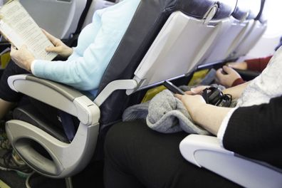 In this real situation a row of unidentifiable passengers are sitting in their seats on an airplane. The seating is tight. Their legs are touching the seats in front of them.