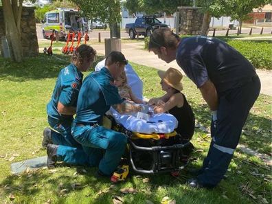 Toddler on a stretcher in a park after being bitten by a snake, surrounded by paramedics and her mum.