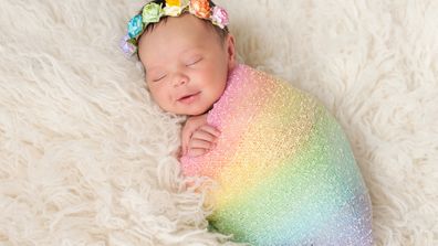 A smiling nine day old newborn baby girl bundled up in a rainbow colored swaddle. She is lying on a cream colored flokati (sheepskin) rug and wearing a crown made of roses.
