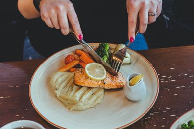 A woman sitting at the table in a cafe, eating redfish steak and mashed potatoes with vegetables on a white plate