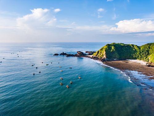 Una lussureggiante collina verde incontra le limpide acque blu dell'oceano. È visibile il litorale roccioso, ornato da rocce di varia forma e dimensione. In lontananza, un cielo nuvoloso dipinge uno sfondo sereno. Scatto effettuato a Lombok.