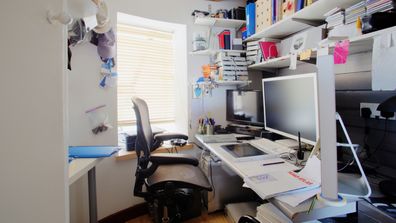 A shot of a messy desk in a home office, the room is small and cluttered.