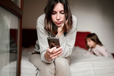 woman work online on mobile with toddler kid at home. tired woman sitting on the bad in a bedroom with smartphone in her hands and trying to work.