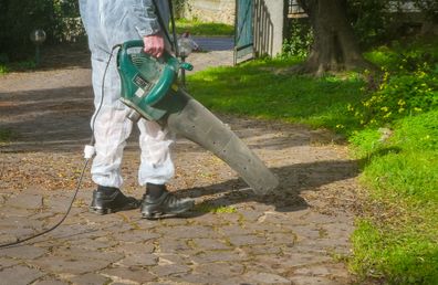 Pria yang mengenakan setelan putih menggunakan blower daun di taman, di hari yang cerah