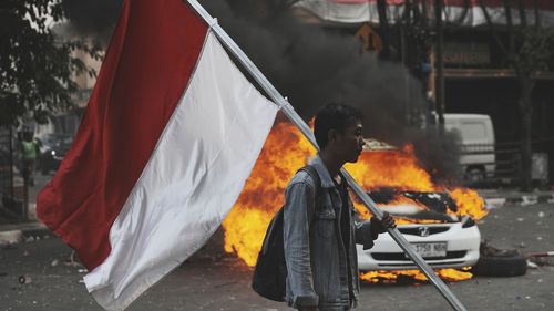 Um homem carrega uma bandeira nacional indonésia perto de um carro em chamas durante um protesto após a morte de um piloto de entrega em confrontos entre a polícia de Riot e os estudantes que protestam contra contra os legisladores, em Jacarta, Indonésia, sexta -feira, 29 de agosto de 2025 (AP Photo/Achmad Ibrahim)