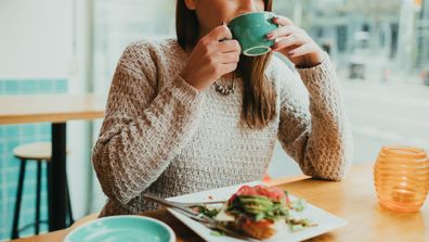 Young woman sitting in the coffee bar, drinking coffee from a blue mug and eating healthy vegan avocado toast for breakfast