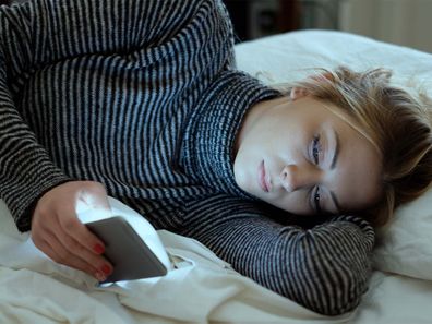 Teenage girl lying on her side looking sadly at her phone.