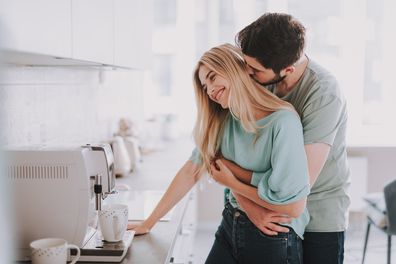 Bearded guy standing behind blonde girl and kissing her. They are waiting for hot drink