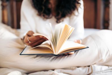 Unrecognizable young woman turning the pages of a book while reading in bed covered with the duvet