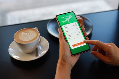 Over the shoulder view of a young Asian woman using a smartphone application to book flight tickets and plan her holiday while having breakfast in a cafe