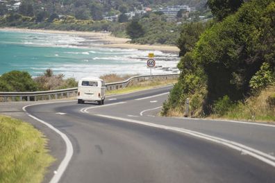 A Volkswagen Kombi (or Transporter) van driving down Great Ocean Road, with Lorne Beach in the background.