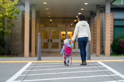 Rear view of a cheerful young mother holding her toddler daughter's hand while walking at crosswalk to the child's school.