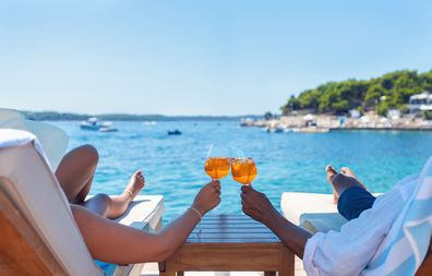 Couple relaxing and toasting with a Aperol spritz cocktail on a beach deck over the ocean. Turquoise ocean is crystal clear.  horizon in the distance.